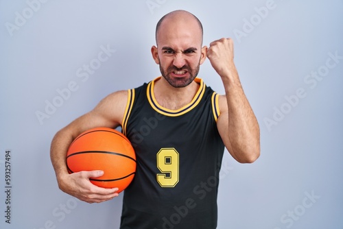 Young bald man with beard wearing basketball uniform holding ball angry and mad raising fist frustrated and furious while shouting with anger. rage and aggressive concept.