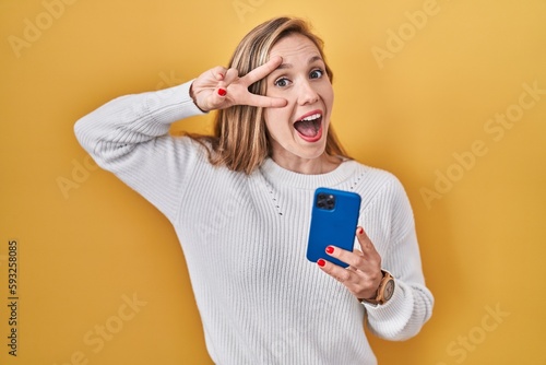 Konstfotografi Young blonde woman using smartphone typing message doing peace symbol with finge