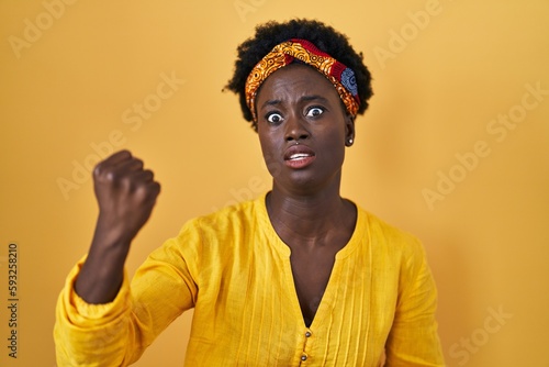 African young woman wearing african turban angry and mad raising fist frustrated and furious while shouting with anger. rage and aggressive concept.