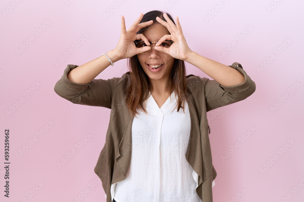 Middle age chinese woman wearing glasses over pink background doing ok gesture like binoculars sticking tongue out, eyes looking through fingers. crazy expression.