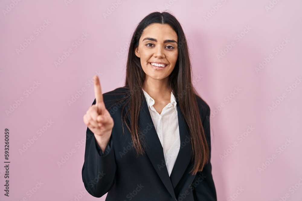 Young brunette woman wearing business style over pink background showing and pointing up with finger number one while smiling confident and happy. Young brunette woman wearing business style over pink background showing and pointing up with finger number one while smiling confident and happy.