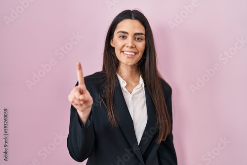 Young brunette woman wearing business style over pink background showing and pointing up with finger number one while smiling confident and happy.