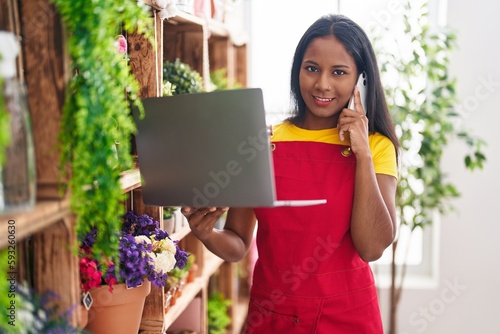 Young beautiful woman florist talking on smartphone using laptop at florist