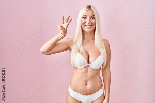 Caucasian woman wearing lingerie over pink background showing and pointing up with fingers number three while smiling confident and happy.