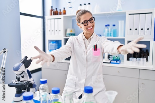 Young woman working at scientist laboratory looking at the camera smiling with open arms for hug. cheerful expression embracing happiness.