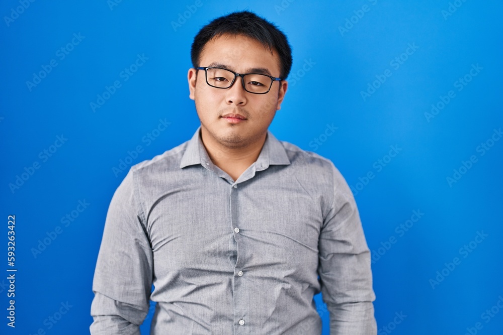 Young chinese man standing over blue background relaxed with serious expression on face. simple and natural looking at the camera. Young chinese man standing over blue background relaxed with serious expression on face. simple and natural looking at the camera.