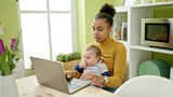 Mother and son sitting on table working online while care baby at dinning room