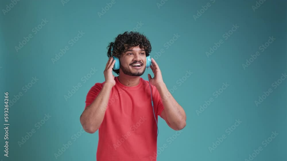Young smiling happy cheerful student Indian man 20s dancing with headphones listen to music have fun in coral t-shirt on blue studio background. People lifestyle concept Young smiling happy cheerful student Indian man 20s dancing with headphones listen to music have fun in coral t-shirt on blue studio background. People lifestyle concept
