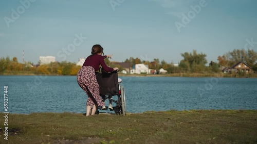 Aunt spends time with niece in wheelchair. Young woman points finger to house on opposite bank of river. Preschooler girl undergoes rehabilitation after injury backside view