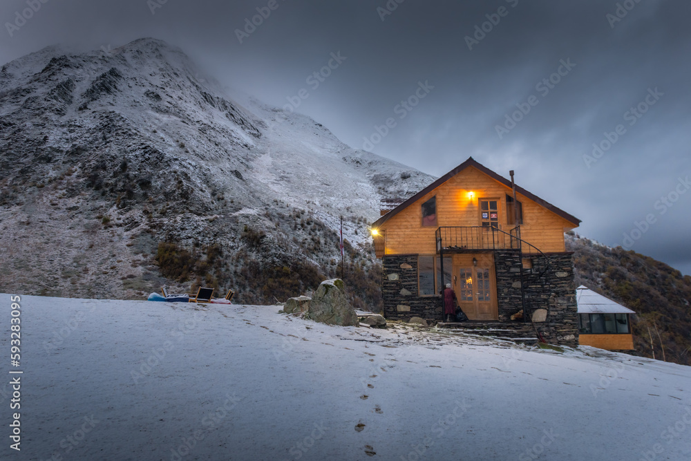Fototapeta premium Timber cottage in the snowy day with a snow mountain background during twilight time