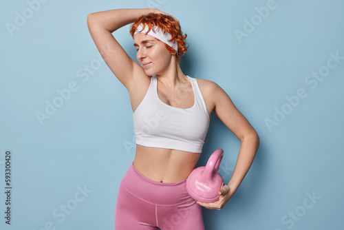 Horizontal shot of sporty motivated woman lfts weight in gym demonstrates strength and motivation dressed in sportswear being in good physical shape isolated over blue background. Healthy lifestyle