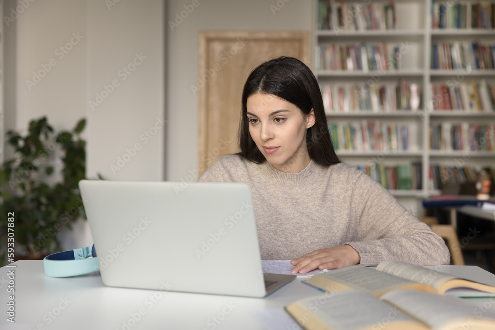 Fototapeta premium Attractive student girl sits at desk in library prepare for university exams or high school admission using laptop looks focused, makes exercise, browse information, do task. E-learning, modern tech