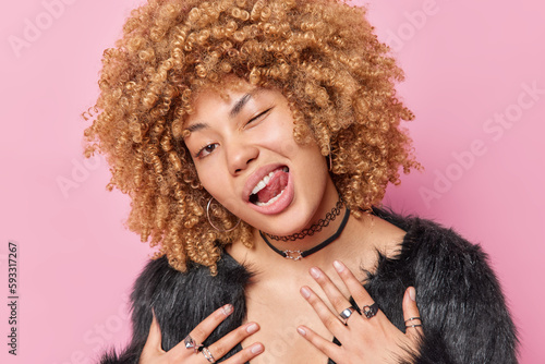 Headshot of curly haired beautiful young woman winks eye and sticks out tongue has cheeky facial expression wears fur coat and chain poses against pink background. Women and fashion concept. photo