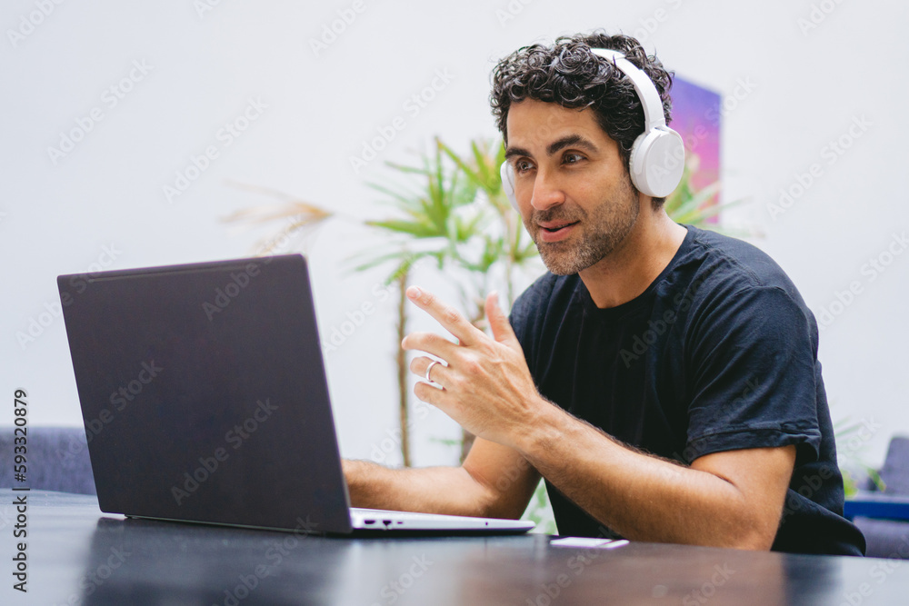 Middle-aged Latin man making a video call on his laptop while wearing a wireless headphone. Middle-aged Latin man making a video call on his laptop while wearing a wireless headphone.