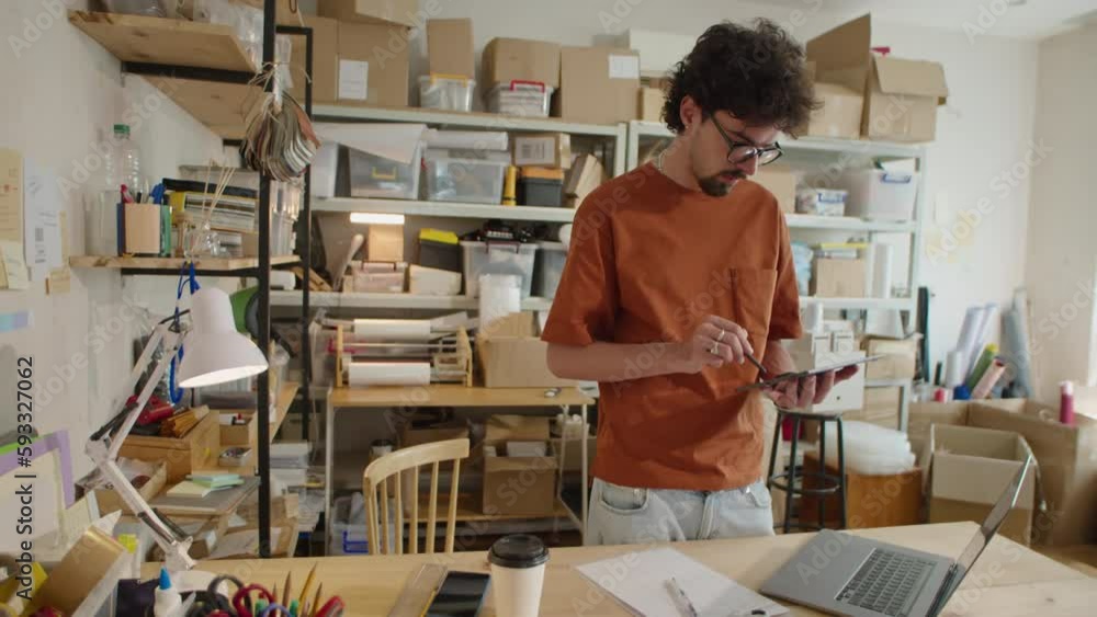 Medium long shot of young male worker using digital tablet while standing beside desk in warehouse of delivery service company Medium long shot of young male worker using digital tablet while standing beside desk in warehouse of delivery service company