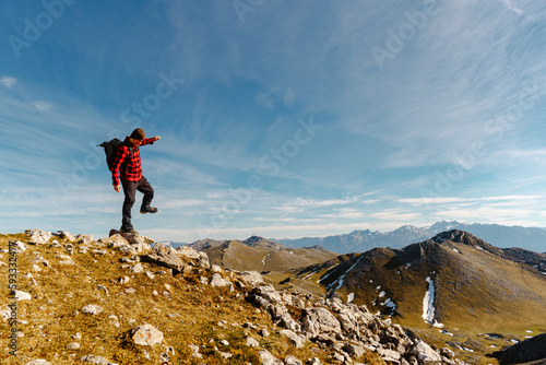 white male mountaineer with red clothes and backpack balancing on a rock on a mountain peak. lone hiker hiking a mountain trail on a sunny day. sport and adventure.