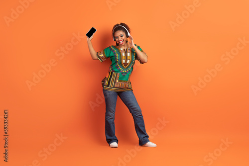 Joyful black woman in african clothes using headphones and smartphone