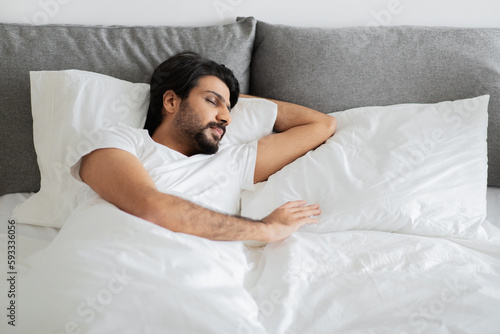 Young middle eastern guy in white t-shirt sleeps, lies on bed with empty space in bedroom interior, top view