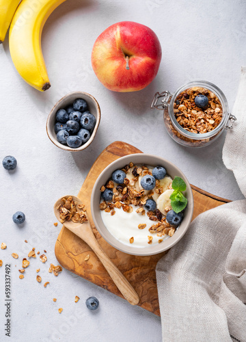 Homemade baked granola with yogurt and blueberries in a bowl on a light background with fresh berries, banana and apple. Healthy vegetarian breakfast with muesli.