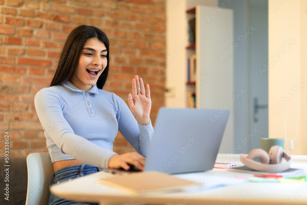 Excited spanish lady having video call on laptop, waving hand, saying hi in living room interior, free space Excited spanish lady having video call on laptop, waving hand, saying hi in living room interior, free space