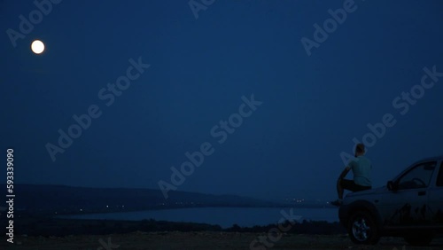 Man sitting alone on the hood of his car by a river and staring into the distance under a night sky photo