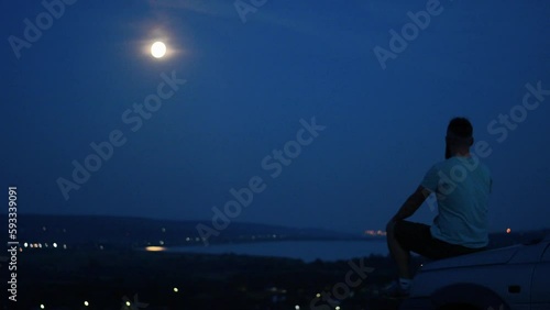 Man sitting alone on the hood of his car by a river and staring into the distance under a night sky photo