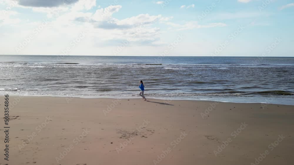 Young girl on sandy coast. Girl leaves footprints on sand. Attractive woman in blue sweater on beach. Girl walking along seashore. Aerial view of seashore. Beautiful ocean waves. Beach with sand Young girl on sandy coast. Girl leaves footprints on sand. Attractive woman in blue sweater on beach. Girl walking along seashore. Aerial view of seashore. Beautiful ocean waves. Beach with sand