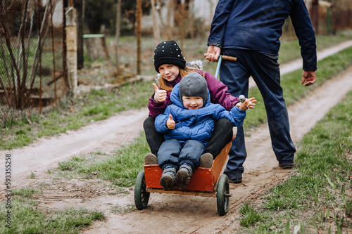 A strong adult man, a father drives, carries on a cart along the road in the village, the countryside, a beautiful smiling, happy girl, a child and a little boy, a son. Photography, portrait.