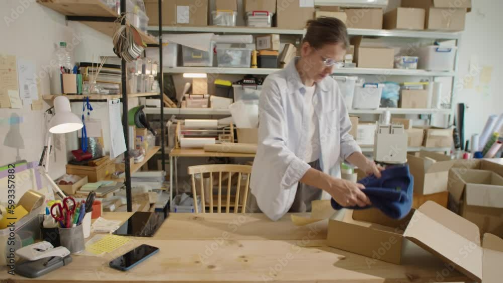 Woman packing clothes into cardboard parcel box while preparing order for shipping during workday in online store warehouse Woman packing clothes into cardboard parcel box while preparing order for shipping during workday in online store warehouse