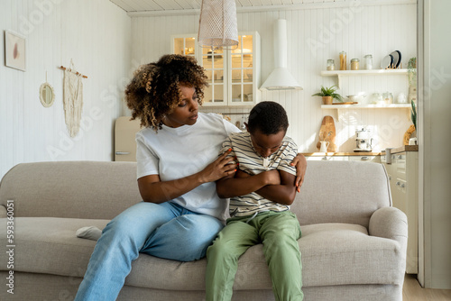 Loving african american mother embracing hugging sad little son supporting child, mom parent trying to make peace with offended upset kid, sitting together on sofa at home. Parent-child conflict photo