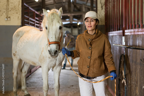 Female stable worker leads a white horse out of the stall