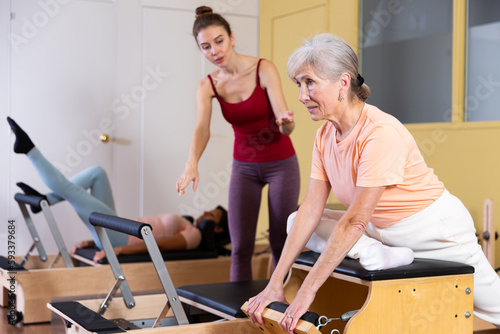 Fototapeta Naklejka Na Ścianę i Meble -  Mature European woman practicing Pilates in a group workout with a female instructor trains on a combined chair in a ..fitness studio