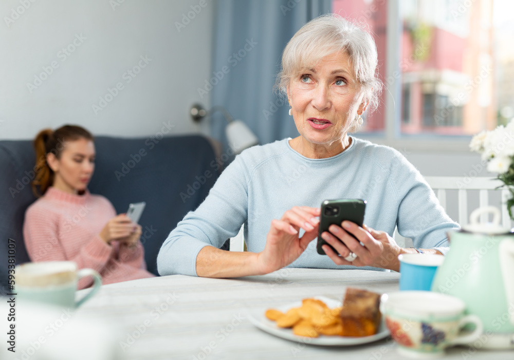 Modern positive elderly woman sitting at table in cozy dining room, using smartphone to chat with friends.