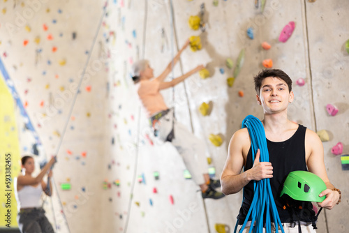 Smiling active young guy standing with rope on shoulders and helmet in hand against artificial training climbing wall in adventure park