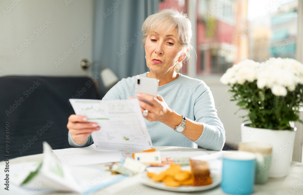 Focused senior woman reading documents at home table and using cell phone to make copy of papers.