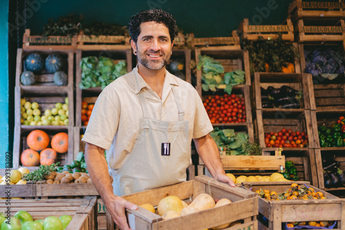 фотографія Middle-aged Latin man with a smile holding a crate of squash in a greengrocer's shop while looking at the camera