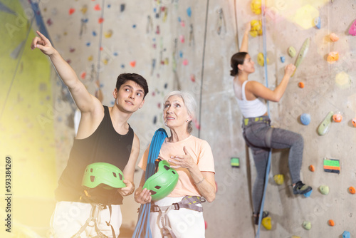 Young man and mature woman standing with blue safety belts and helmet before practicing rock-climbing on climbing wall in adventure park