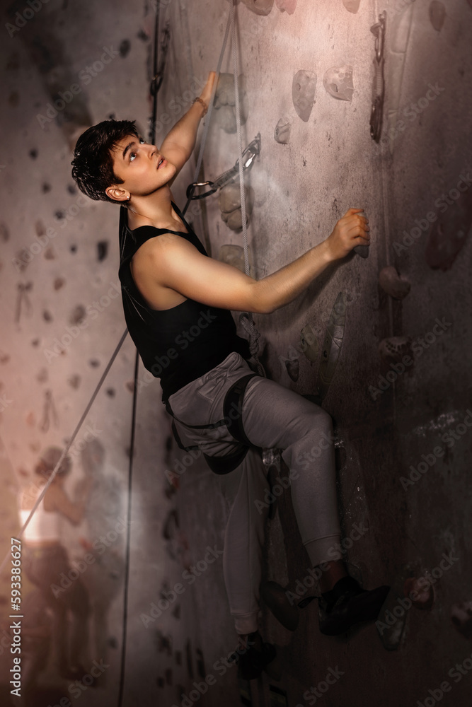 Positive extreme young guy with rope training on concrete climbing rock wall to maintain healthy discipline in indoor adventure park