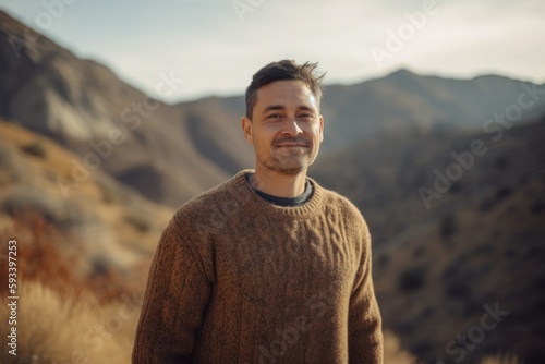 Handsome young man in warm sweater looking at camera and smiling while standing in the mountains