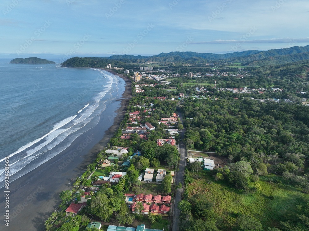 Jaco Beach, a popular surfing destination in Costa Rica, is captured in stunning aerial shots from a drone, showcasing its long sandy shore, waves, palm trees, and colorful beachfront buildings.