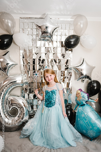 The girl is dressed in a blue dress of Elsa. Happy child celebrating a birthday party 6 years. Photo wall, photo zone decoration white, black, silver balloons. Idea decor an arch in luxury style. photo