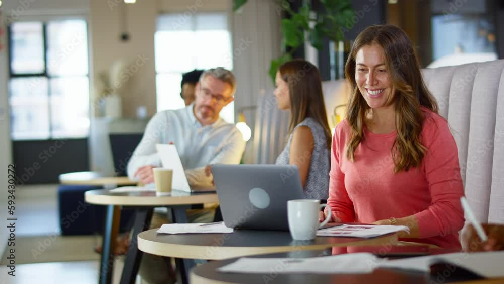 Mature businesswoman making video call on laptop in informal seating area of modern office on coffee break - shot in slow motion Mature businesswoman making video call on laptop in informal seating area of modern office on coffee break - shot in slow motion