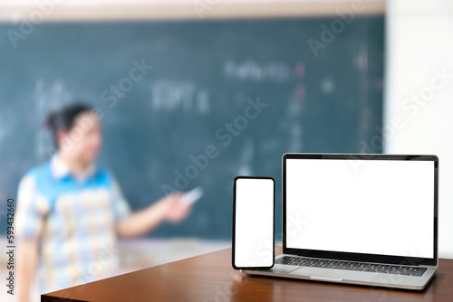front view Blank screen of black digital laptop and smartphone on wooden table, teacher standing in front of classroom. educational software website technology