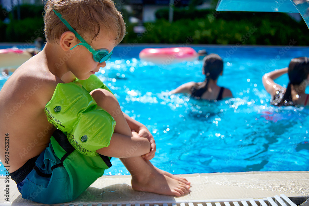 Frustrated boy in bathing shorts and glasses is sitting on the curb by