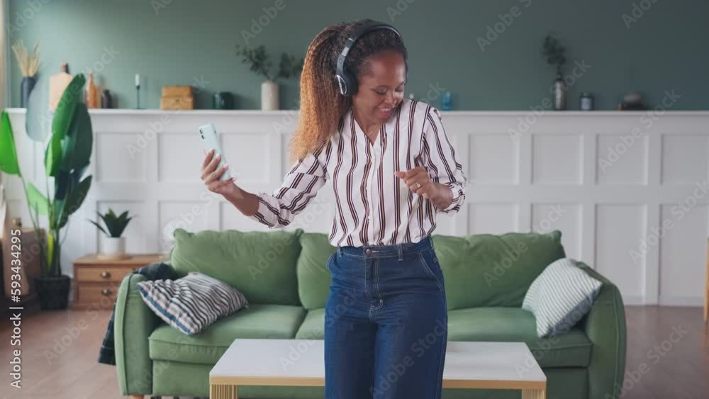 Young happy elegant African American woman in headphones dancing talking on video call through phone enjoying online communication with boyfriend located in spacious bright living room Young happy elegant African American woman in headphones dancing talking on video call through phone enjoying online communication with boyfriend located in spacious bright living room