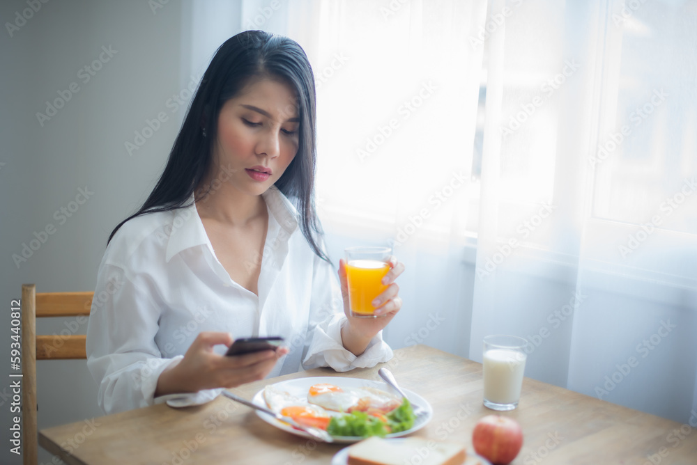 Beautiful asian woman eyes look on the screen through emails and messages on her phone as she picked up a glass of juice and breakfast on the table.