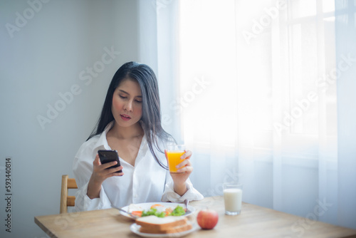 Beautiful asian woman looking at the phone in her hand while about to drink juice and breakfast on the table.