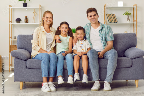 Young happy Caucasian family with two children watching TV together sitting on sofa in living room. Little girl points remote control at camera while sitting next to her mom, brother and dad.