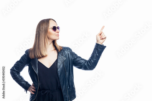 Young stylish woman points up with her index finger. Pretty woman in black leather jacket and sunglasses. Isolated on white background. Space for text.
