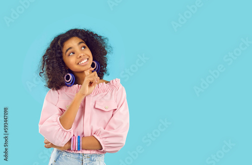 Happy child thinking about something good. Cute kid imagining future vacation and smiling. African American girl in trendy outfit dreaming of summer trip and looking up on blue copy space background